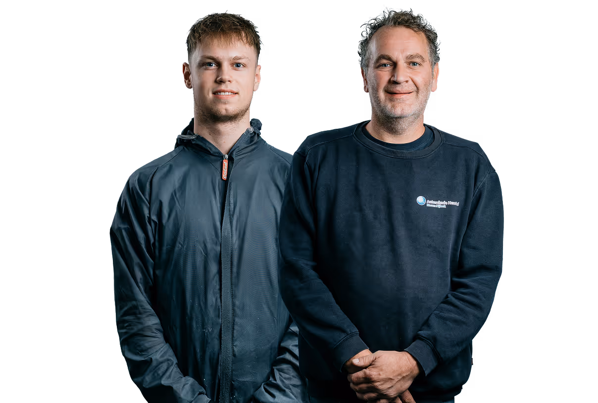 Twee mannen, een jongere met een zwarte jas links en een oudere met een donkerblauwe trui rechts, glimlachend en voor een zwarte achtergrond.