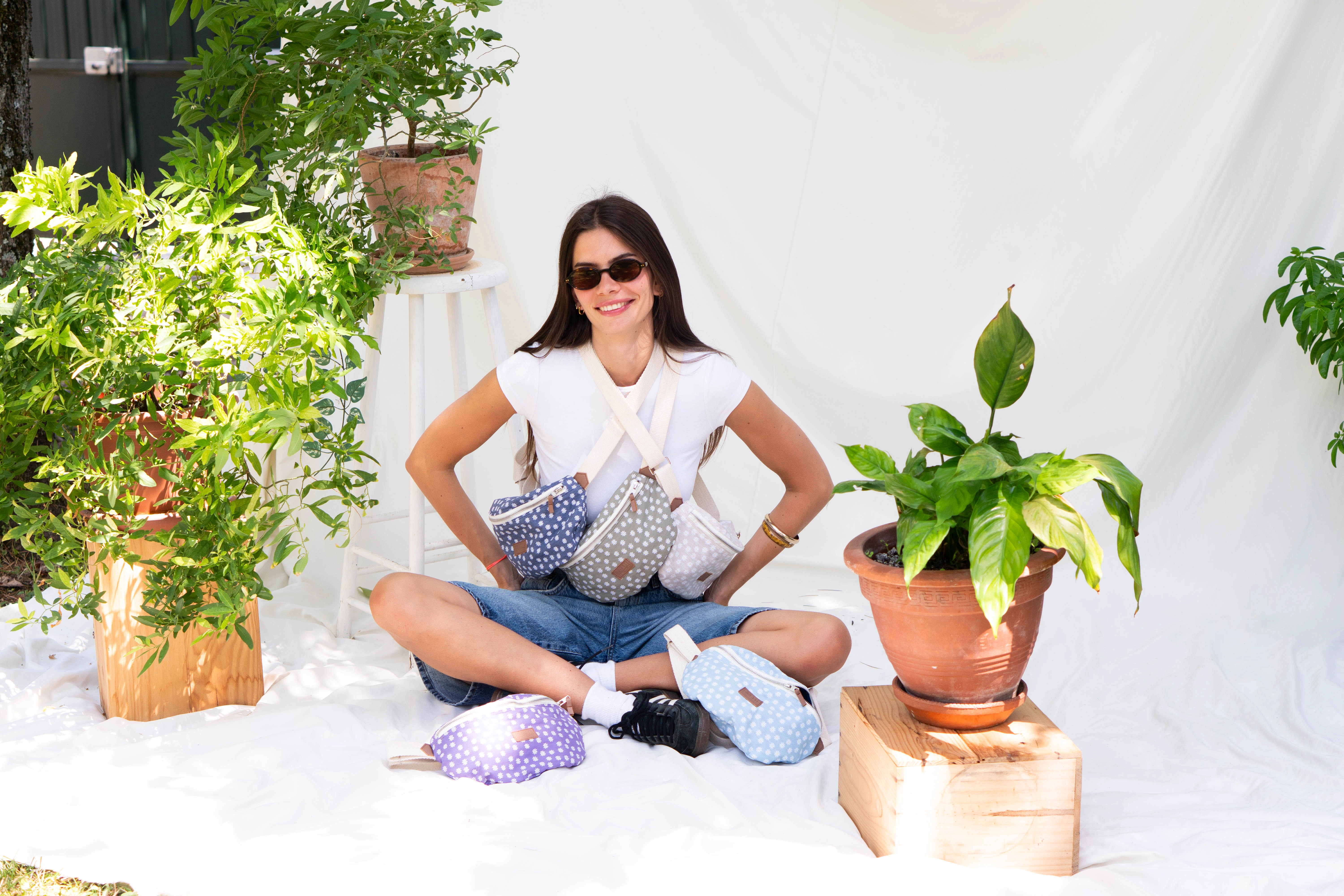 Une femme souriante assise sur un fond blanc décoré avec plusieurs plantes. Elle porte divers accessoires de la marque Binette, dont des bananes.