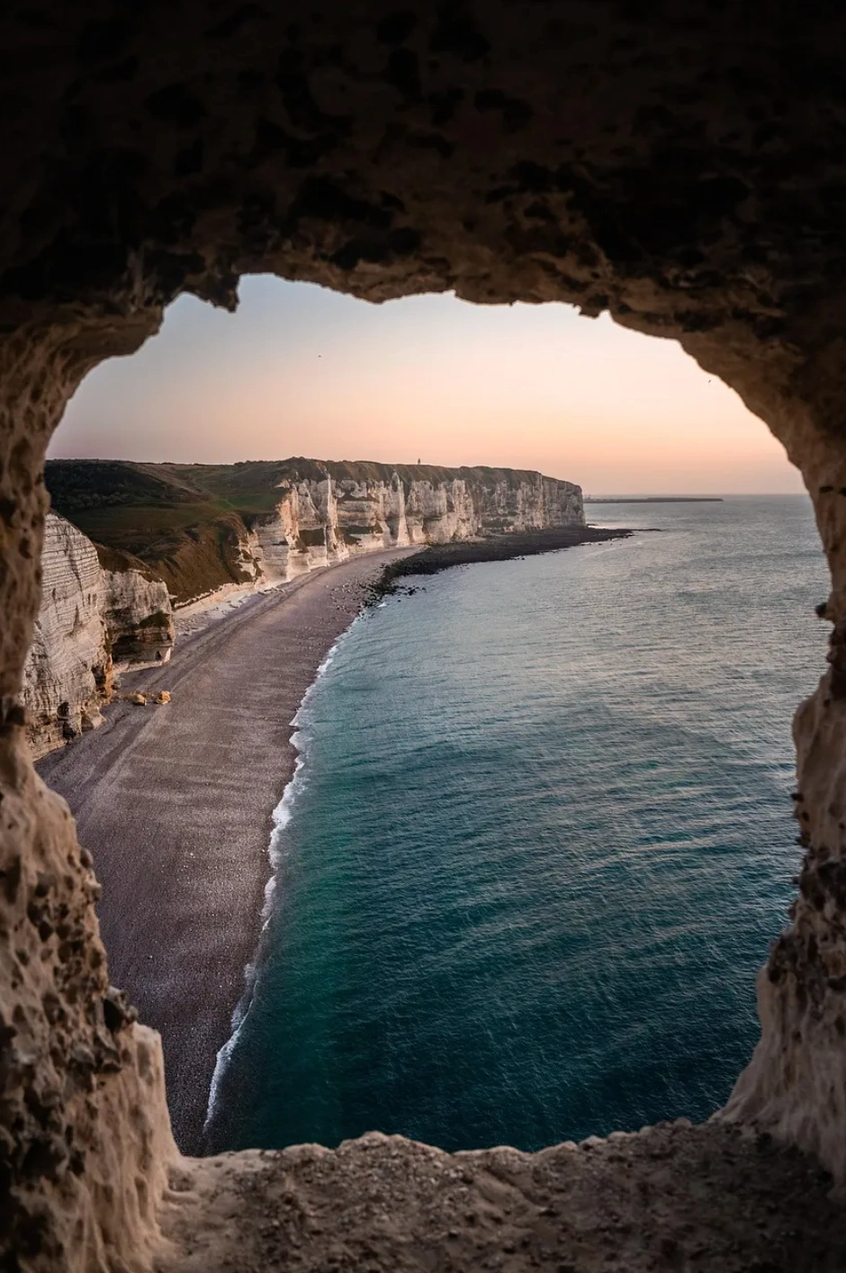 Vue d'une plage et de falaises le long du littoral, encadrées par l'ouverture d'une grotte, au coucher du soleil.