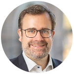 Mann mit kurzen braunen Haaren, Bart und Brille, lächelnd mit einem weißen Hemd und dunklem Blazer.