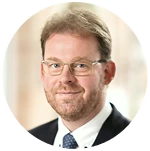 Porträt eines lächelnden Mannes mit Brille, kurzen braunen Haaren und Anzug mit Krawatte.