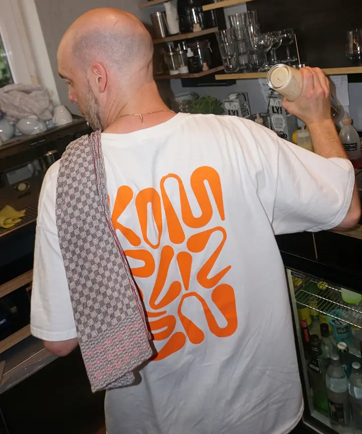Mann mit weißem T-Shirt mit orangefarbenem Muster und Handtuch über der Schulter hält Getränk.