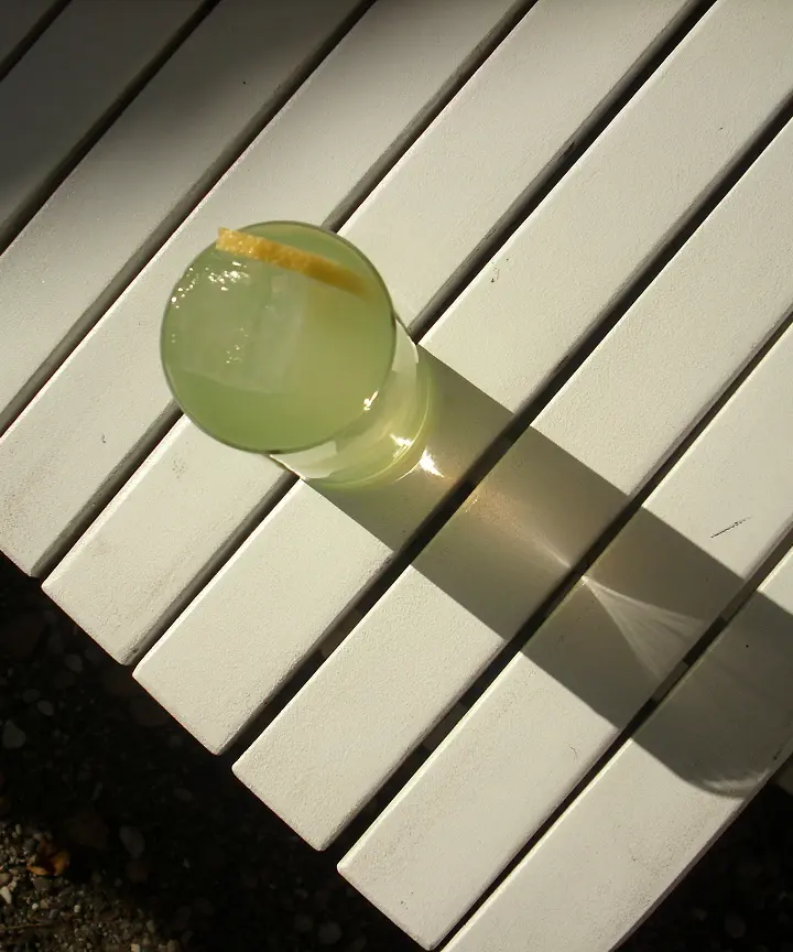 Glas mit Zitronenlimonade und Zitronenscheibe auf weißer Lattenbank im Sonnenlicht.