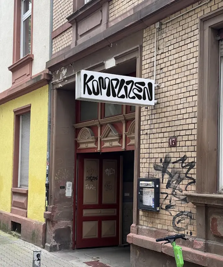 Eingang mit Schild 'Komplizen' an einem Gebäude mit Holz- und Backsteinfassade.