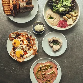 Fünf Teller mit geröstetem Brot, Salat, Humus, Spiegeleier, Aufstrich und Aufschnitt auf Metalltisch.