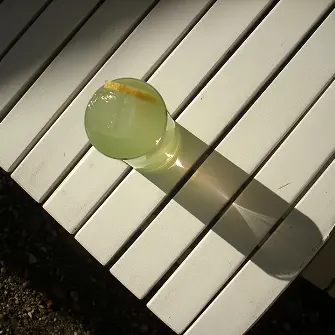 Glas mit Zitronenlimonade und Zitronenscheibe auf weißer Lattenbank im Sonnenlicht.