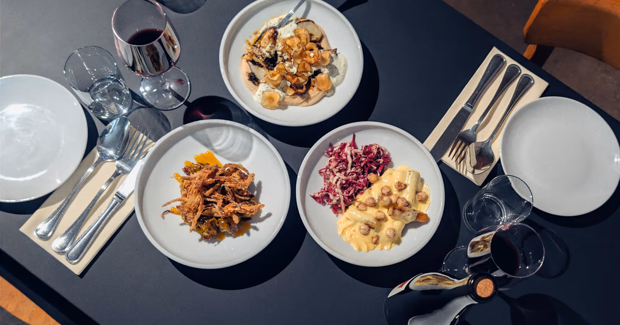Drei Teller mit unterschiedlichem Gourmet-Essen, zwei Gläser Rotwein, leere Teller und Besteck auf dem Tisch.
