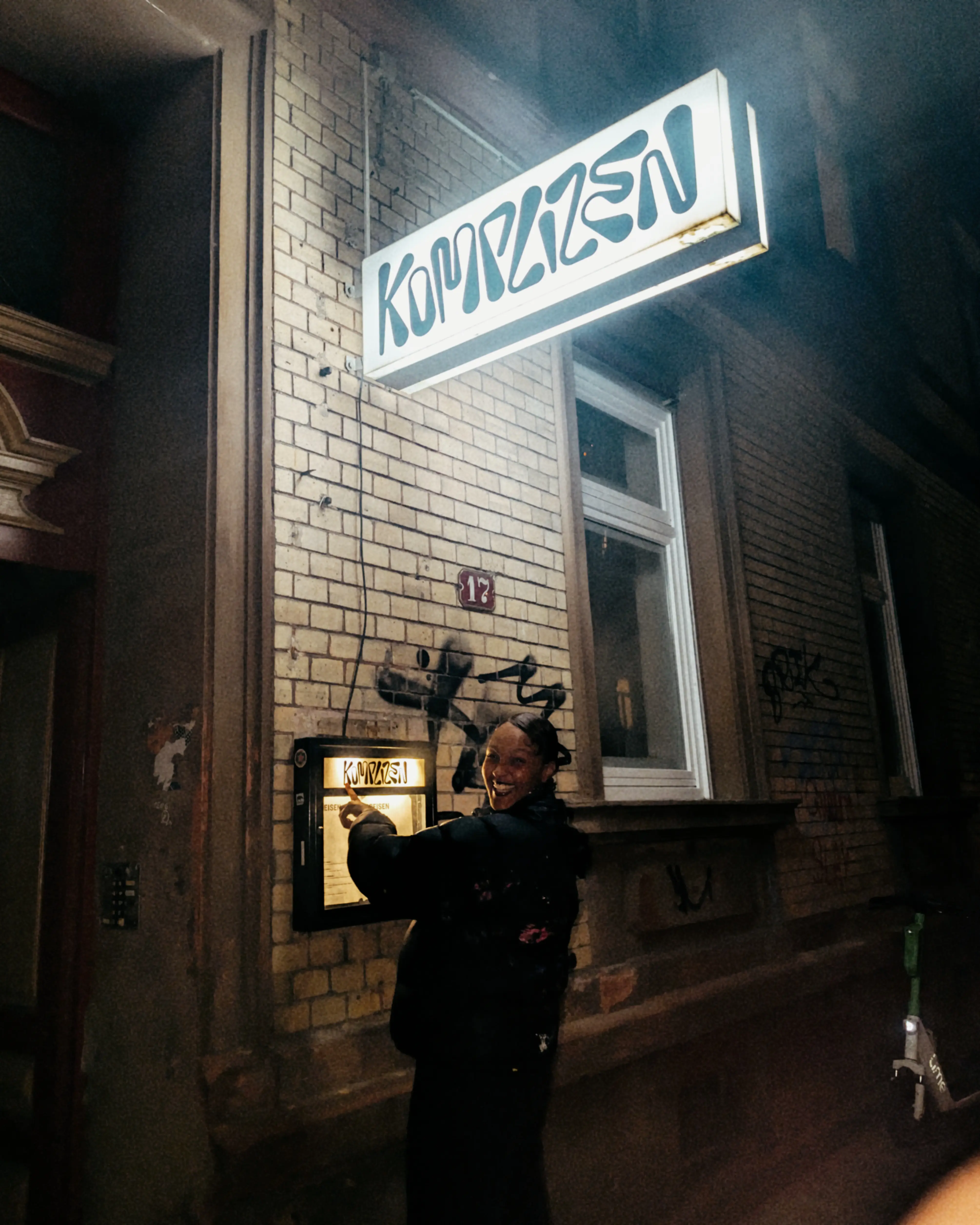 Person zeigt lachend auf ein beleuchtetes Restaurantmenü unter einem Schild mit der Aufschrift Komplizen.