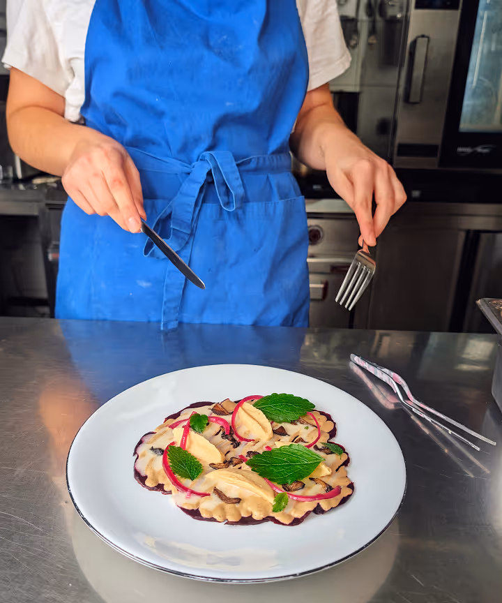 Person mit blauem Schürze hält Messer und Gabel vor Teller mit dekoriertem Ravioli-Gericht.
