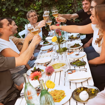 Acht Personen sitzen an einem Tisch im Freien, heben ihre Gläser und lächeln, vor sich stehen Teller mit Essen.