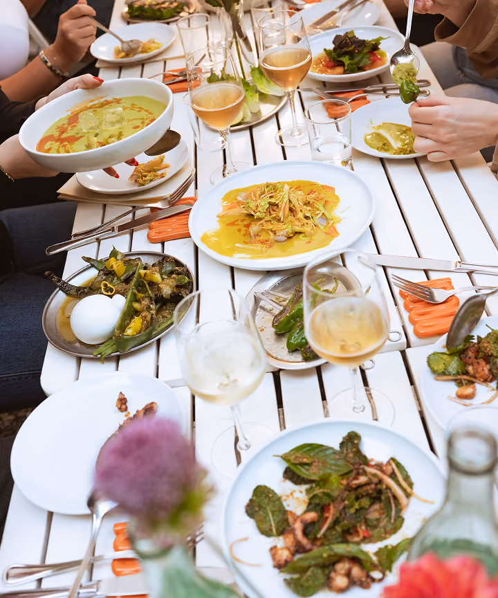 Menschen, die im Freien essen und sich an einem weiß gedeckten Tisch gemeinsam Gerichte und Gläser Weißwein teilen.