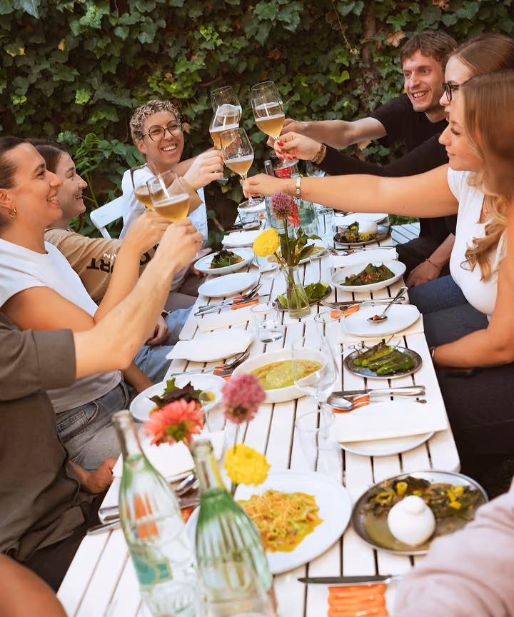 Acht Personen sitzen an einem Tisch im Freien, heben ihre Gläser und lächeln, vor sich stehen Teller mit Essen.