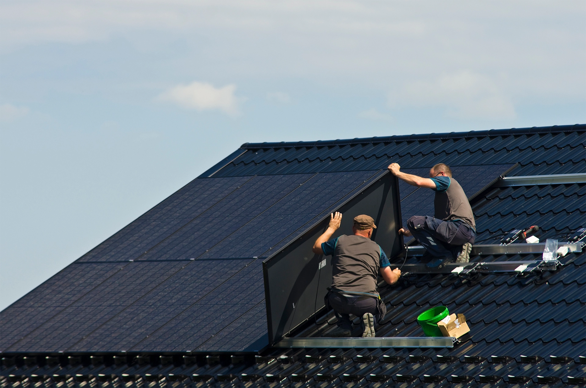 Two workers installing solar panels on a dark tiled rooftop under a clear sky.