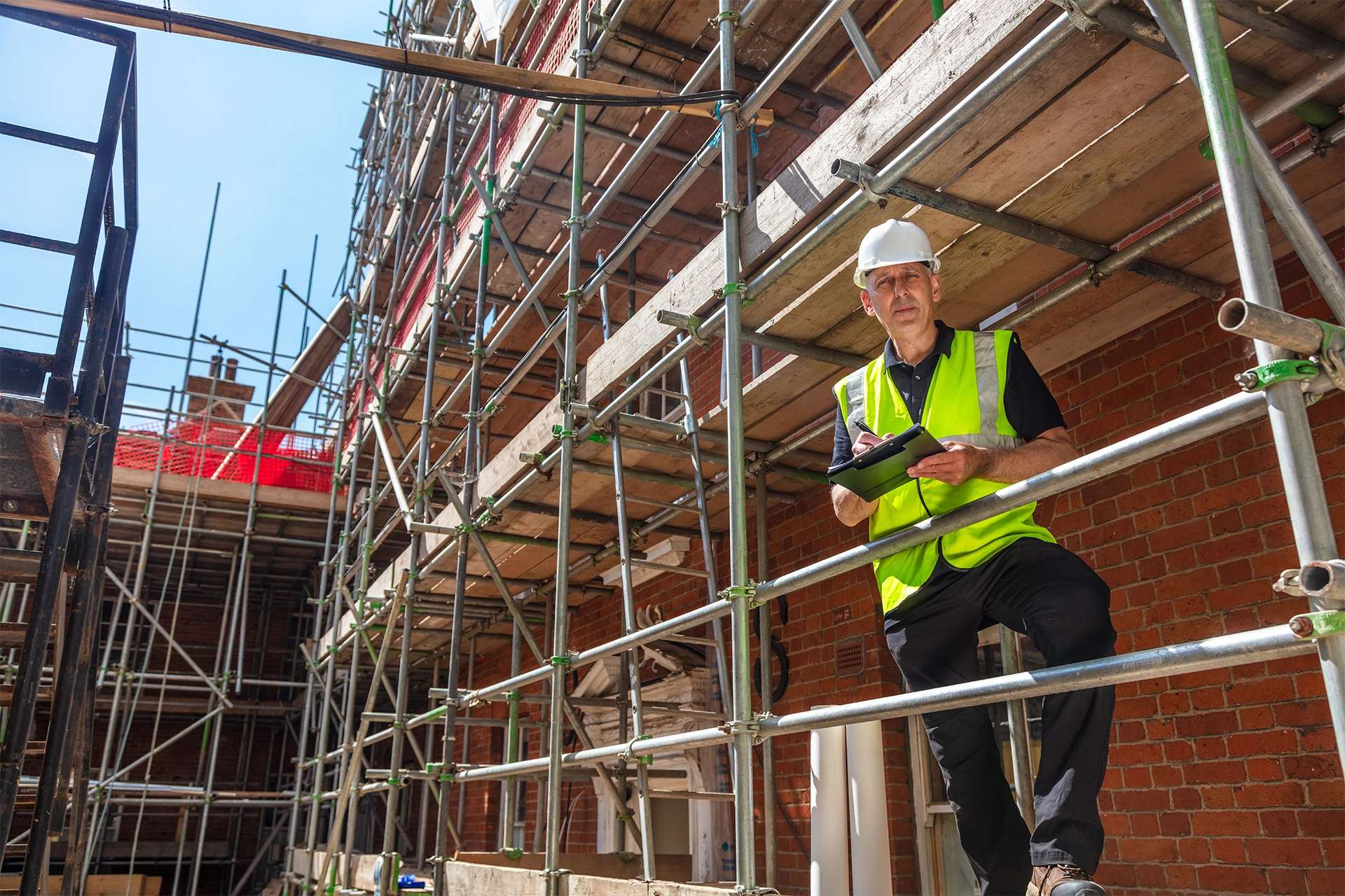 Construction worker in a white helmet and yellow safety vest on scaffolding in front of a brick building holding a clipboard.