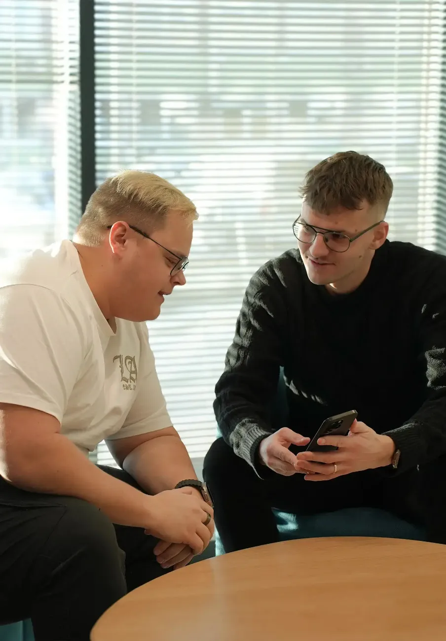 Kaks meest vestlevad, üks hoiab telefoni, teine kuulab tähelepanelikult, mõlemad prillidega ja istuvad laua taga päiksevalguses.