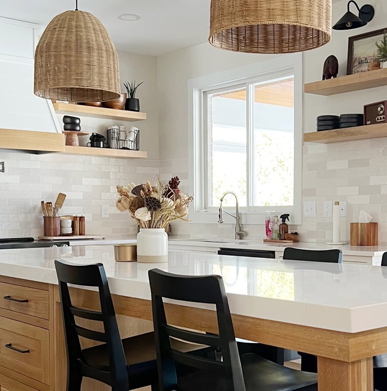 Modern kitchen with light wood cabinets, white tile backsplash, large white island, black chairs, woven pendant lights, and dried flower centerpiece.