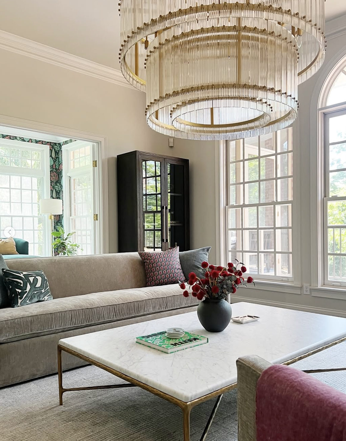 Modern living room with beige sofa, marble coffee table holding a black vase with red flowers, and a large circular chandelier above.