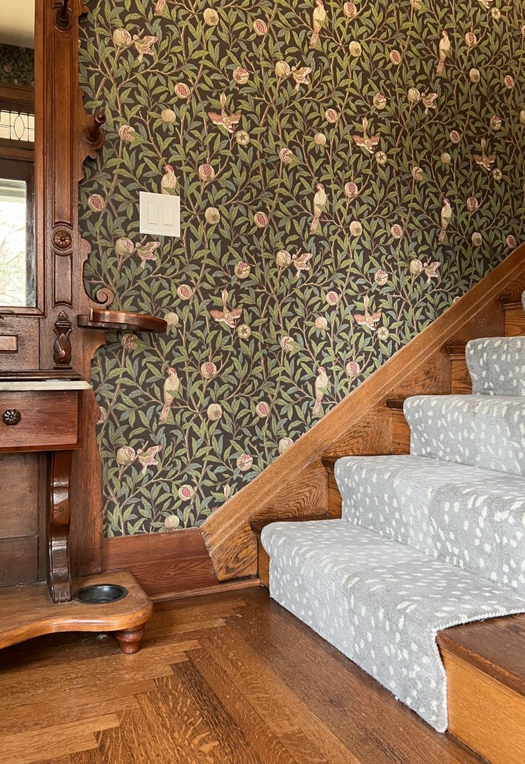 Wooden staircase with light gray patterned carpet runner next to a wall with floral and bird wallpaper, adjacent to a wooden antique furniture piece.