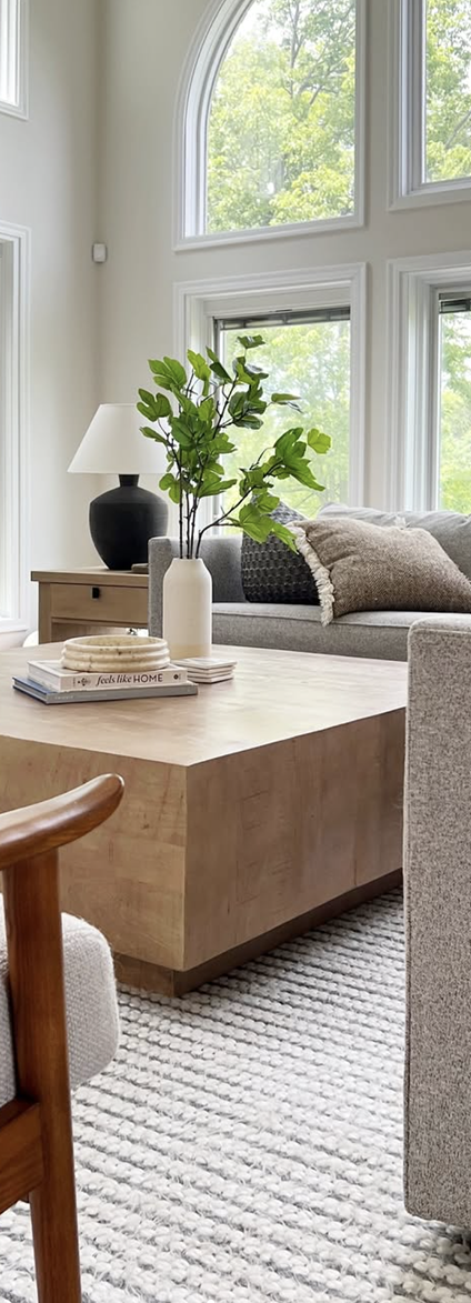 Modern living room with large arched windows, gray sofa with textured pillows, wooden coffee table with a white vase holding green branches, and a lamp on a wooden side table.