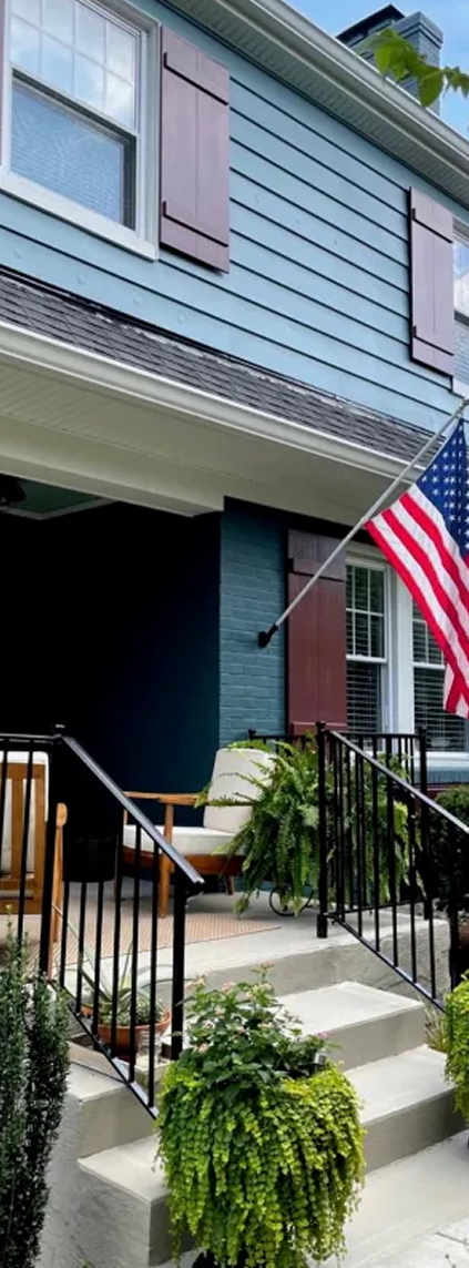 Front porch of a blue house with dark red shutters, black metal railings, potted plants, beige outdoor chairs, and an American flag.