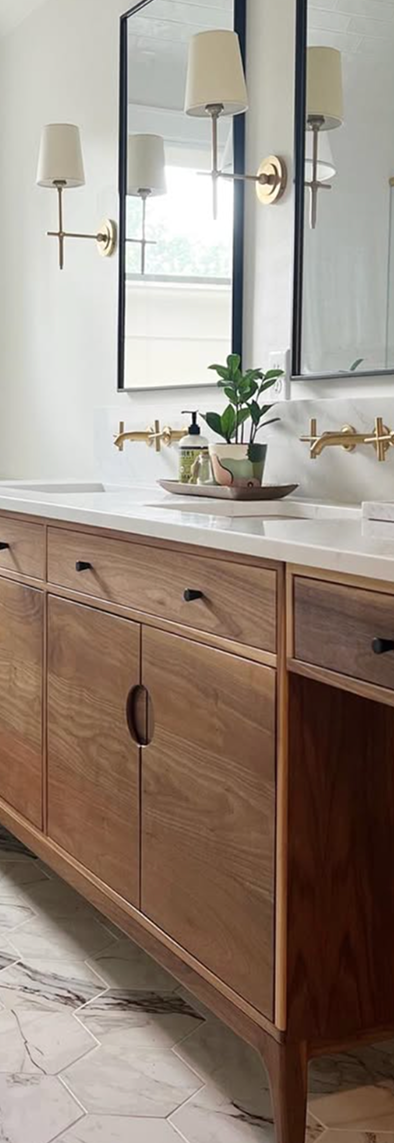 Wooden bathroom vanity with white countertop, gold wall-mounted faucets, oval mirrors, and wall sconces with white lampshades.