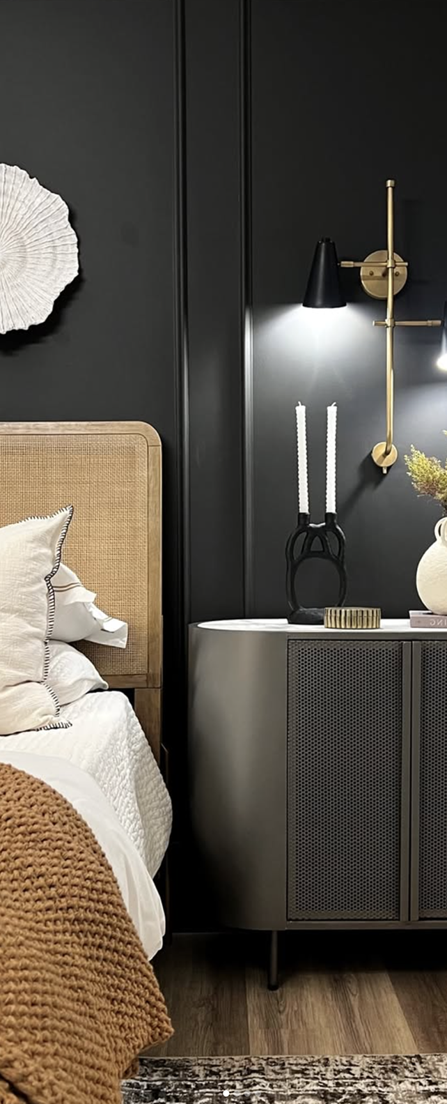 Modern bedroom corner with a beige woven headboard bed, white bedding, a brown knitted throw, black nightstand with decorative candles, a vase with flowers, and a brass wall lamp against a dark gray wall.