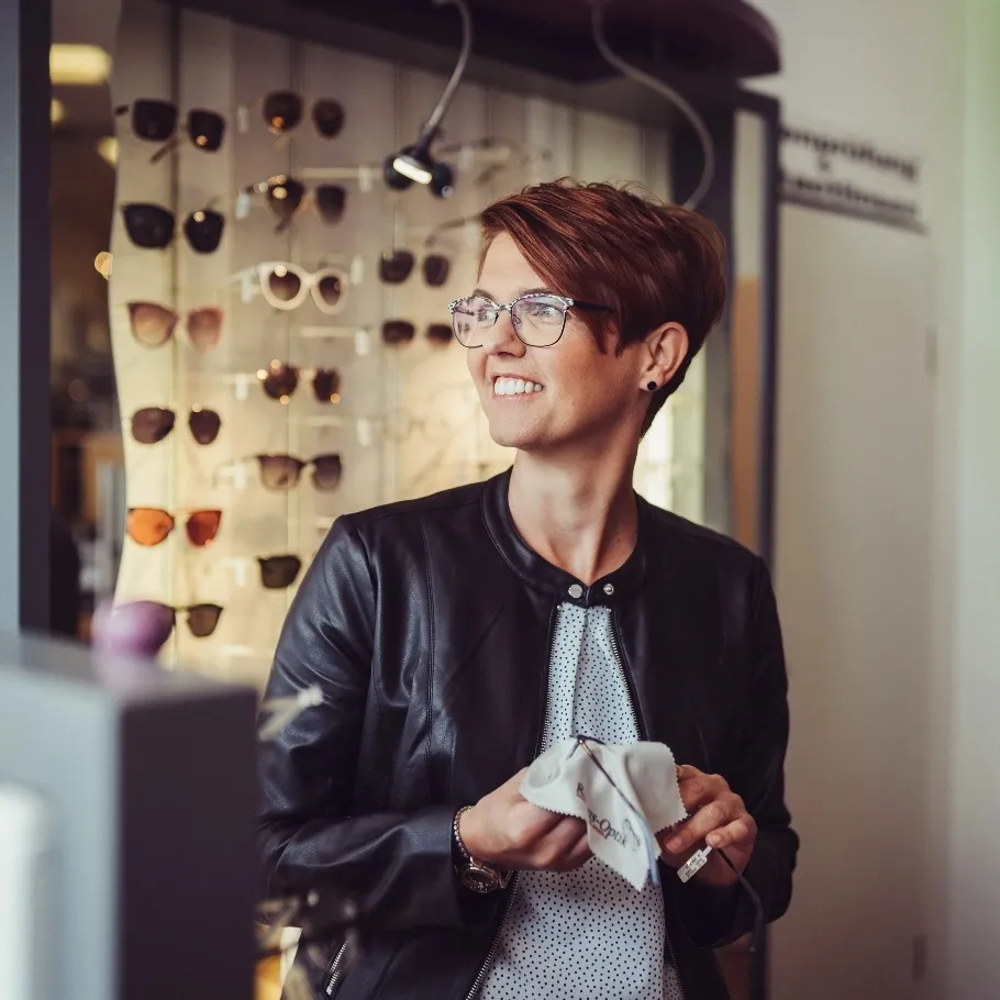 Frau mit kurzen roten Haaren und Brille putzt eine Brille in einem Optikergeschäft mit einer Brillenwand im Hintergrund.