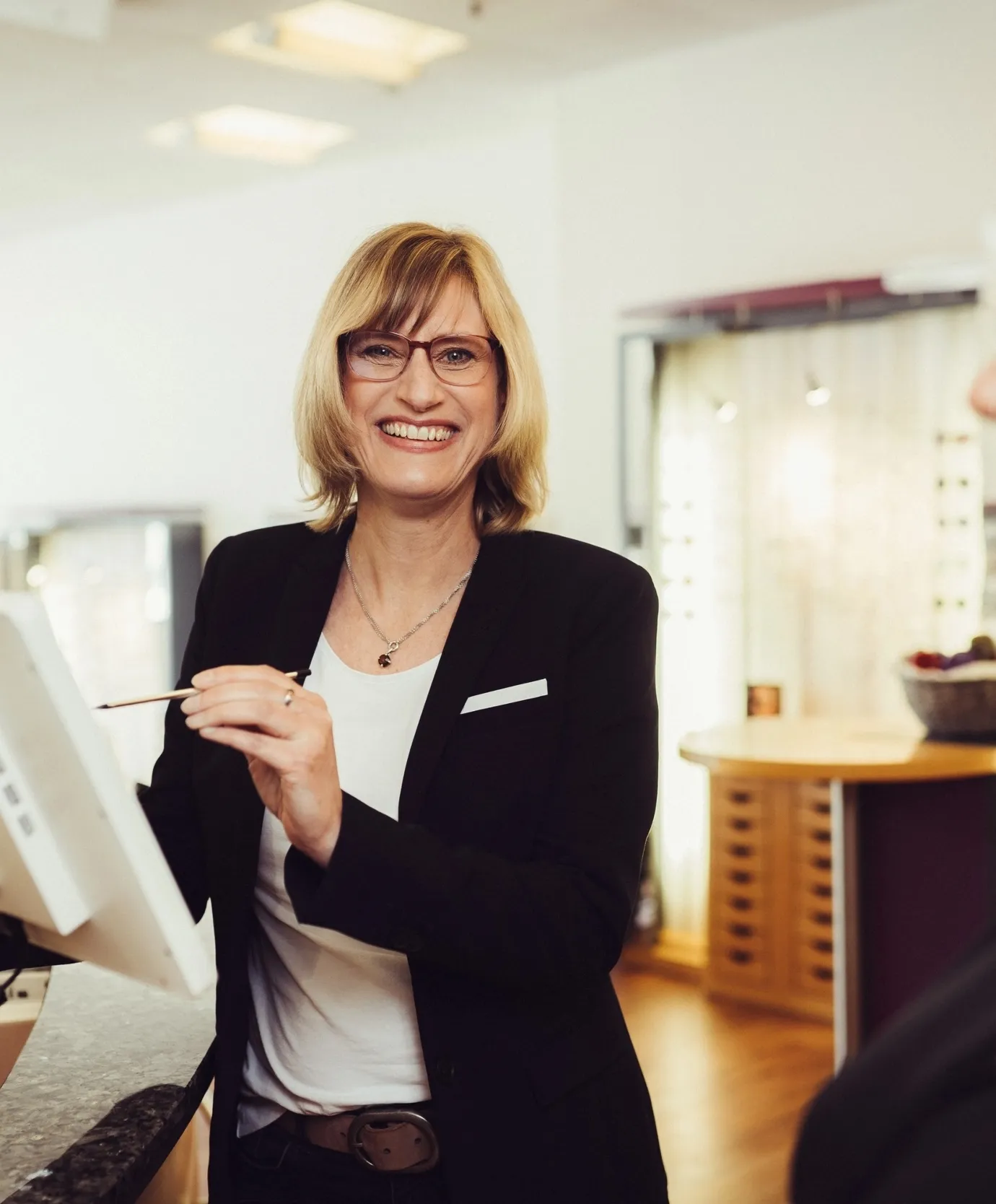 Lächelnde blonde Frau mit Brille in schwarzem Blazer und weißem Shirt vor einem Bildschirm stehend.