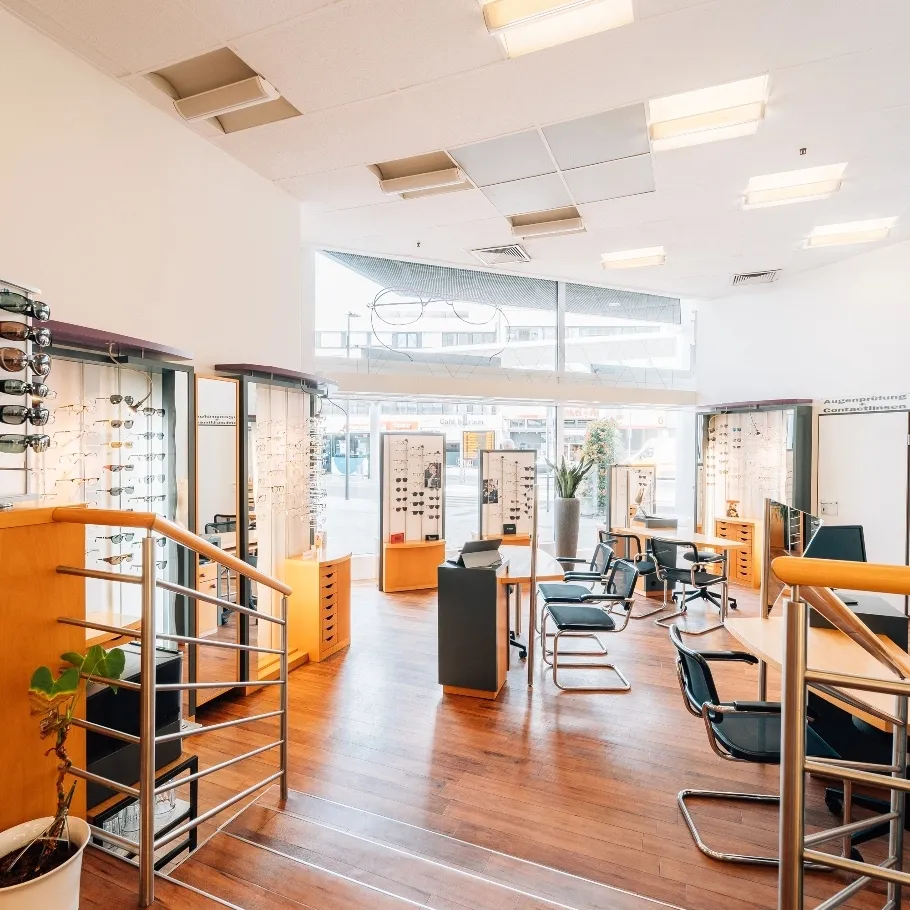 Helle Optikerladen mit Holzboden, mehreren Brillenständern, Stühlen und großen Fenstern mit Blick auf eine Stadtstraße.