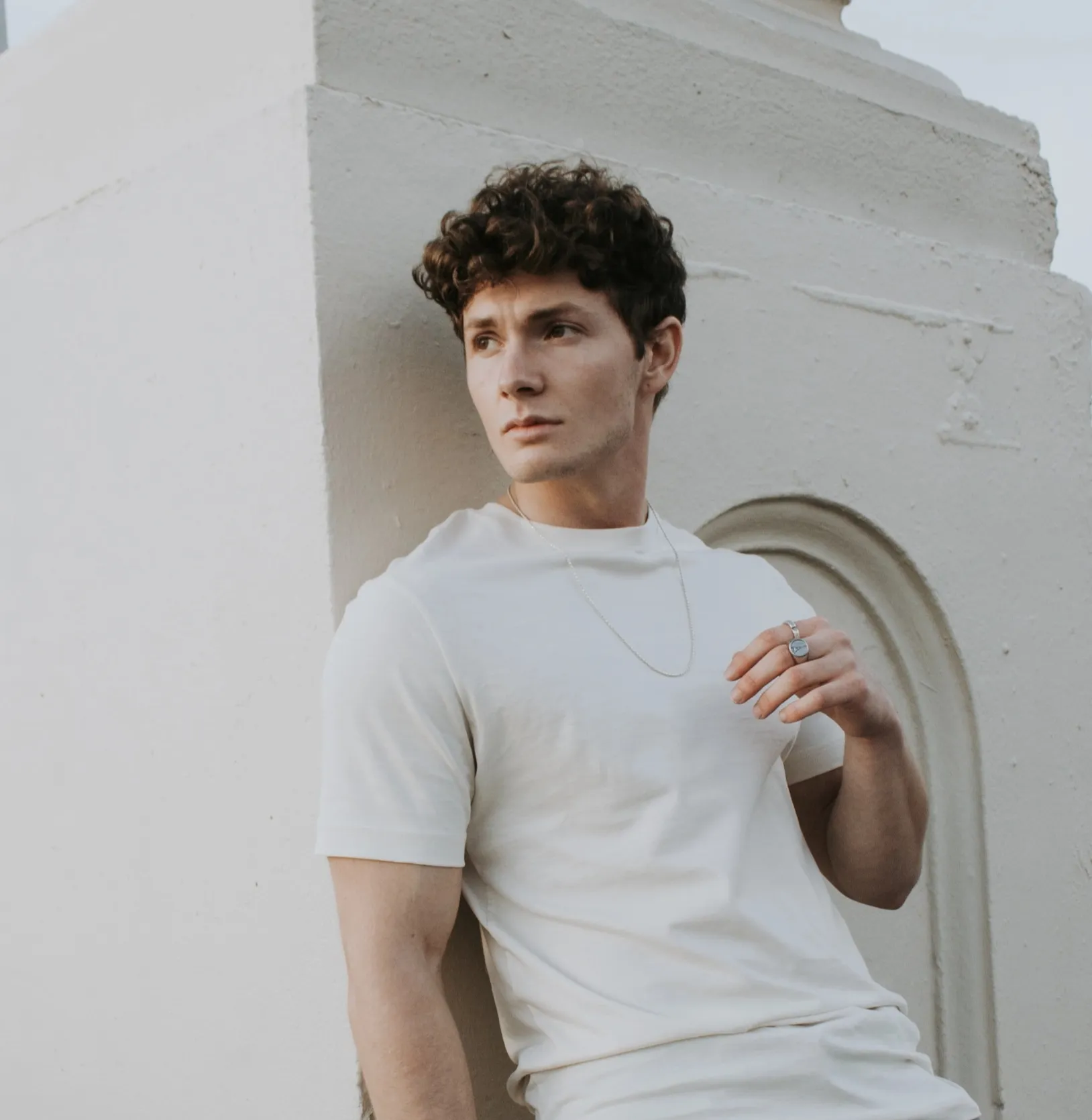 Junger Mann mit lockigem Haar lehnt an einer hellen Wand und schaut nach links.