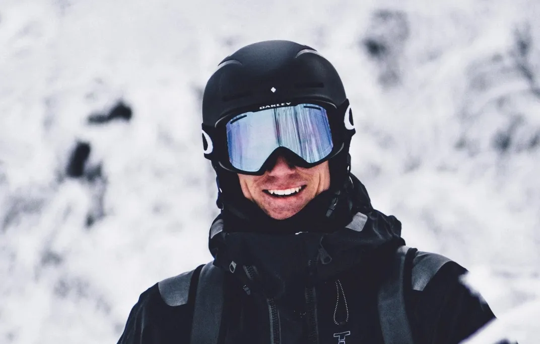 Lächelnder Skifahrer in schwarzer Ausrüstung mit Helm und verspiegelter Skibrille vor schneebedecktem Berg.