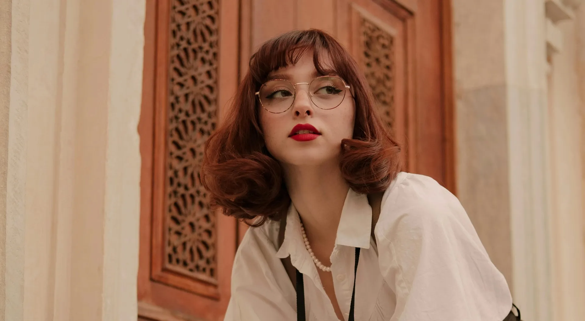 Junge Frau mit roten Lippen, rötlichen gewellten Haaren, runden Brillengläsern und weißem Hemd vor einer Holzverzierung.