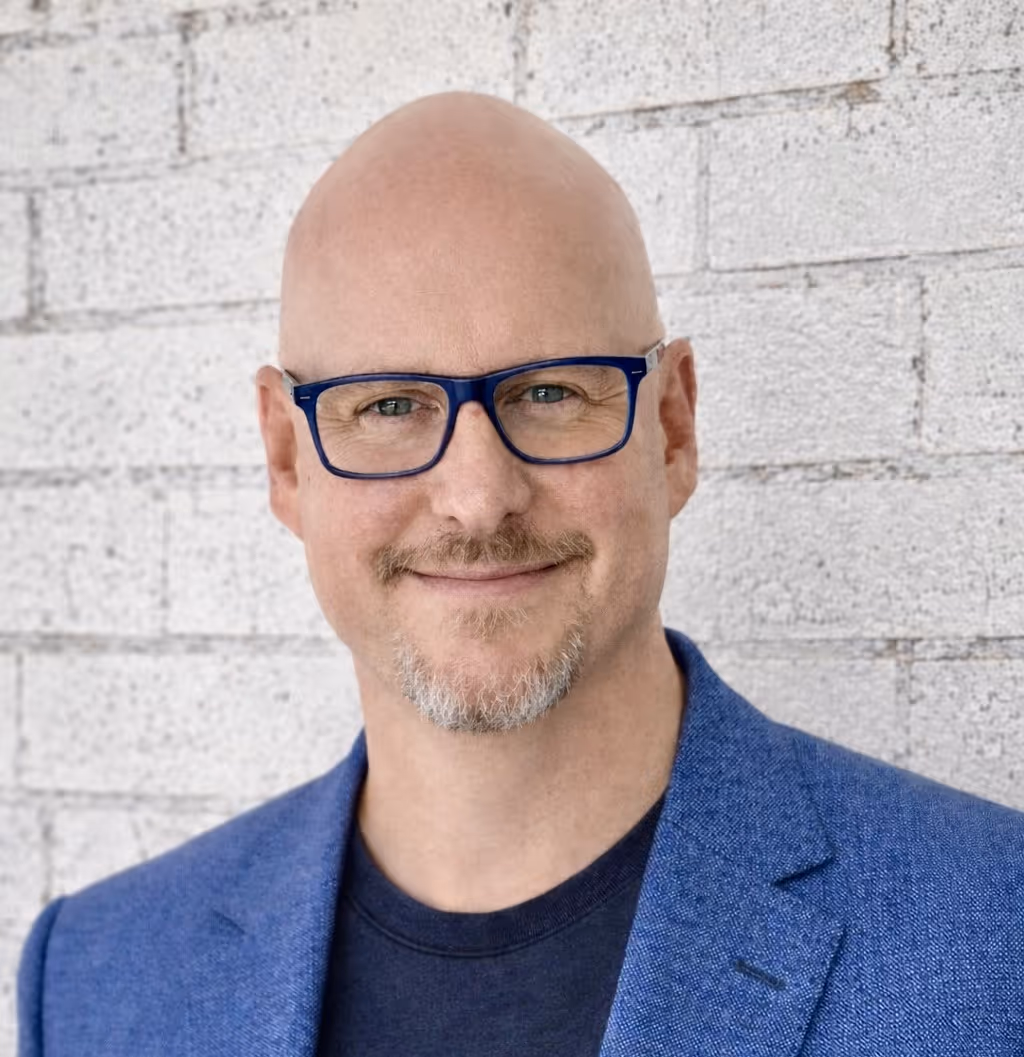 Man in glasses and a blue suit standing against a white brick wall.