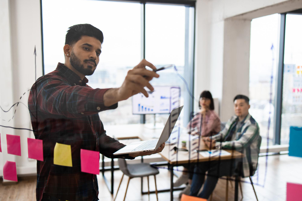 Male presenting notes and graphs written on a glass wall to two people.