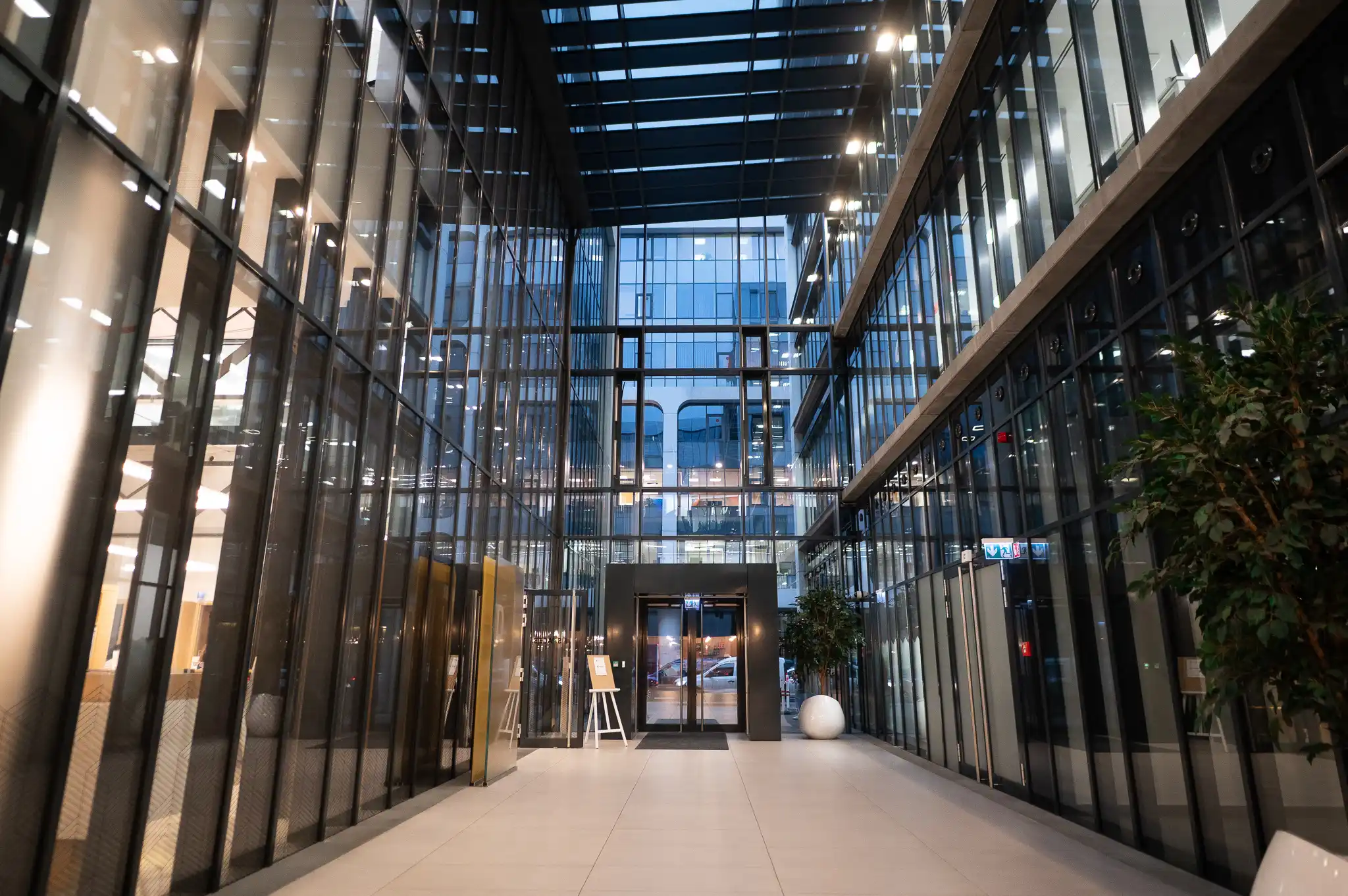 Modern glass-walled atrium lobby with a high skylight ceiling and an entrance doorway at the far end.