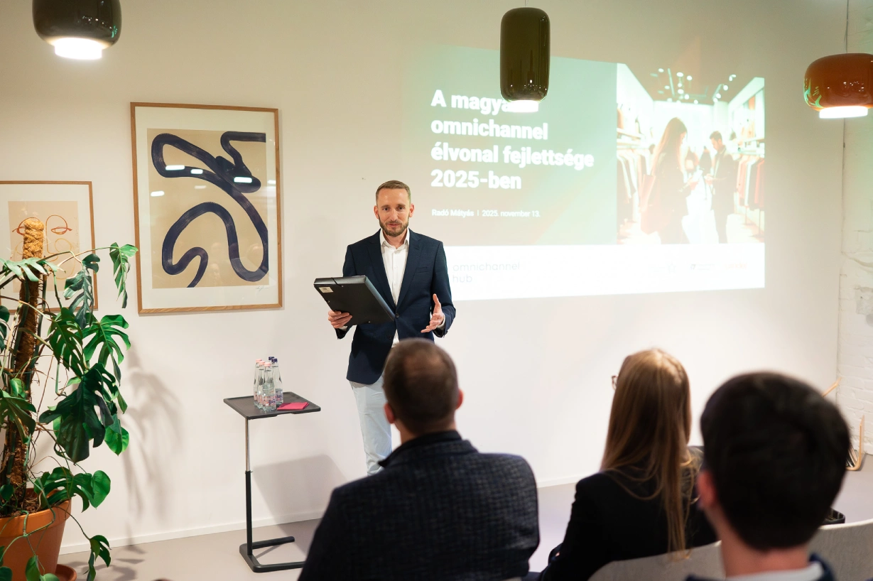Speaker in a suit presents to a small audience, holding a tablet, with a projected slide about Hungary’s omnichannel maturity in 2025 behind him.