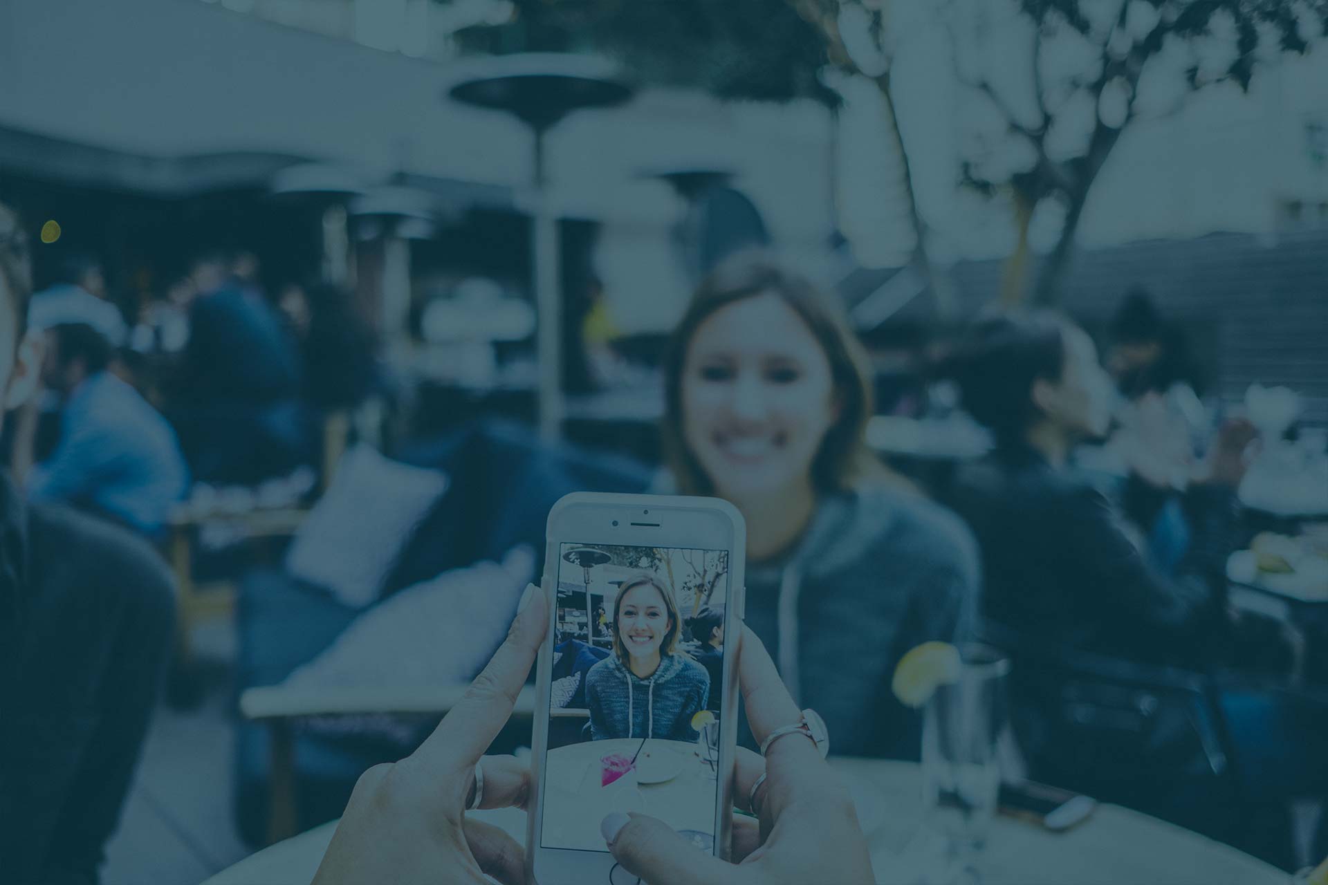 Hands hold a smartphone taking a photo of a smiling woman seated at an outdoor café, with the phone screen in focus and the background blurred.