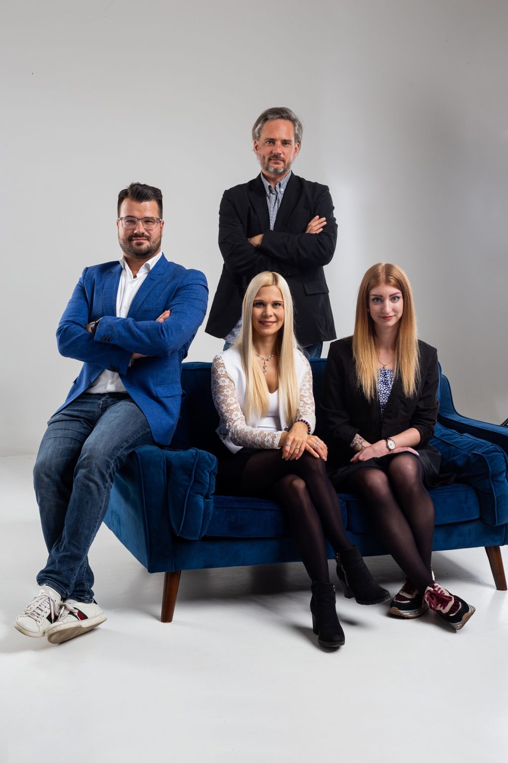 Studio portrait of four business professionals posing with arms crossed; two women sit on a blue velvet sofa while two men stand and sit behind them against a plain gray background.