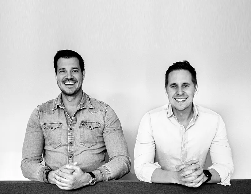 Black-and-white portrait of two smiling men seated side by side at a table, hands clasped, against a plain background.