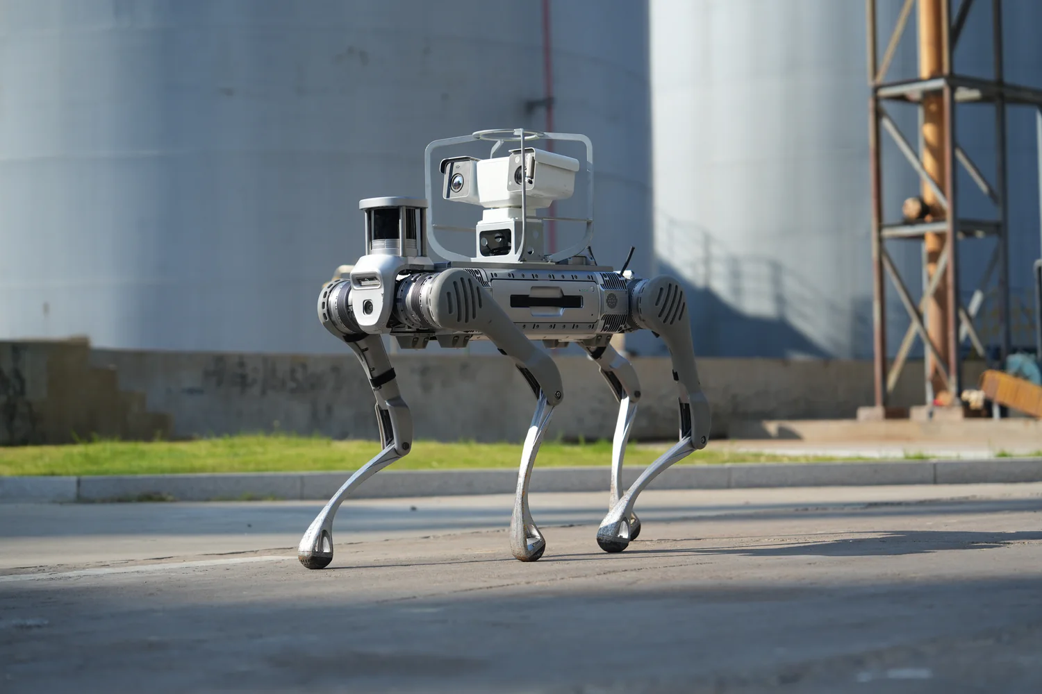 Four-legged inspection robot with mounted cameras walks on a paved surface in an industrial facility, with large storage tanks in the background.