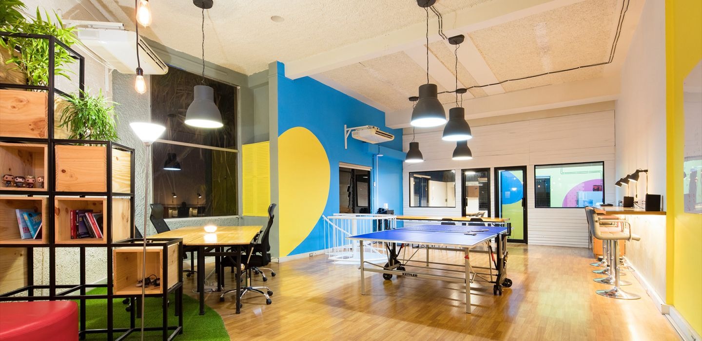 Bright coworking space with a ping-pong table, meeting table, bar-style seating, hanging lights, and colorful wall murals.