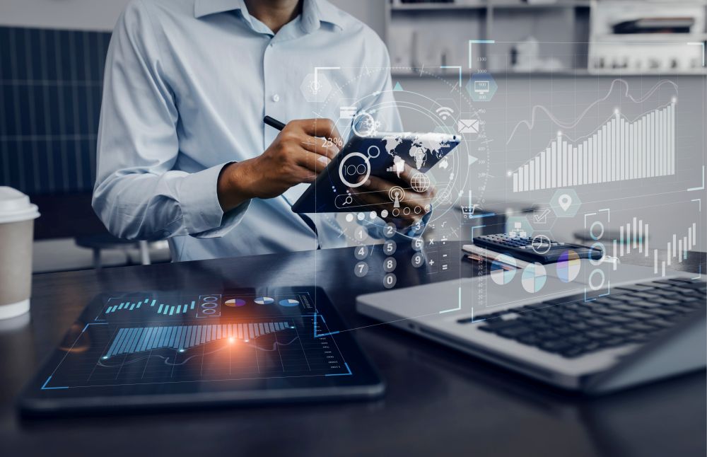 Person uses a tablet at a desk with a laptop and calculator, with transparent data analytics and chart overlays on the image.