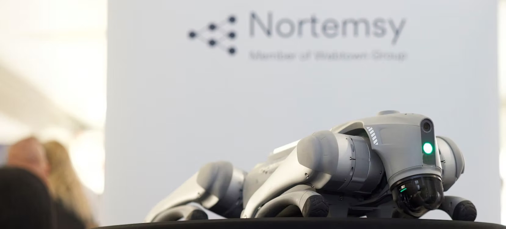 Close-up of a quadruped robot with a glowing status light at a tech event, with “Nortemsy” branding blurred in the background.