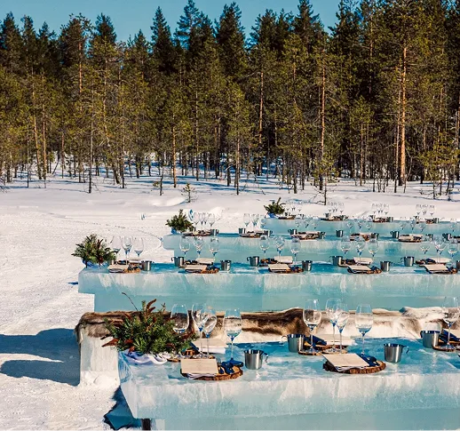 Tables et bancs en glace aménagés en plein air sur la neige avec des couverts, des verres et une décoration de plantes, entourés d'arbres sous un ciel bleu.