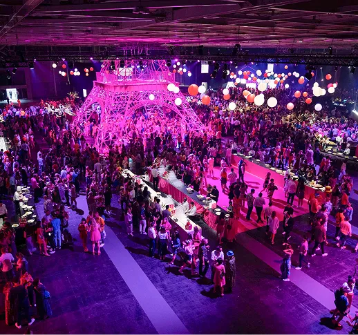 Foule nombreuse autour d'une grande structure illuminée en rose ressemblant à la Tour Eiffel lors d'un évènement en intérieur avec des ballons suspendus.