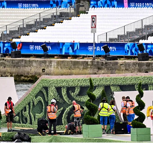 Des techniciens en gilets de sécurité installent une scène décorée avec des arbustes sculptés devant des gradins vides aux Jeux Olympiques de Paris 2024.