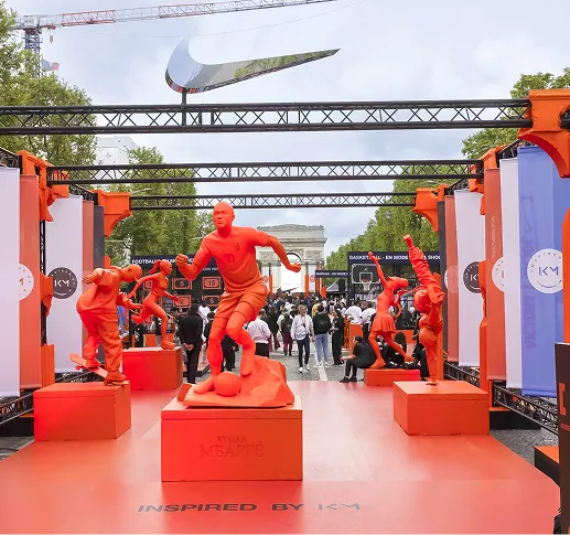 Sculptures of soccer players painted orange displayed on pedestals at an outdoor event with the Arc de Triomphe in the background.