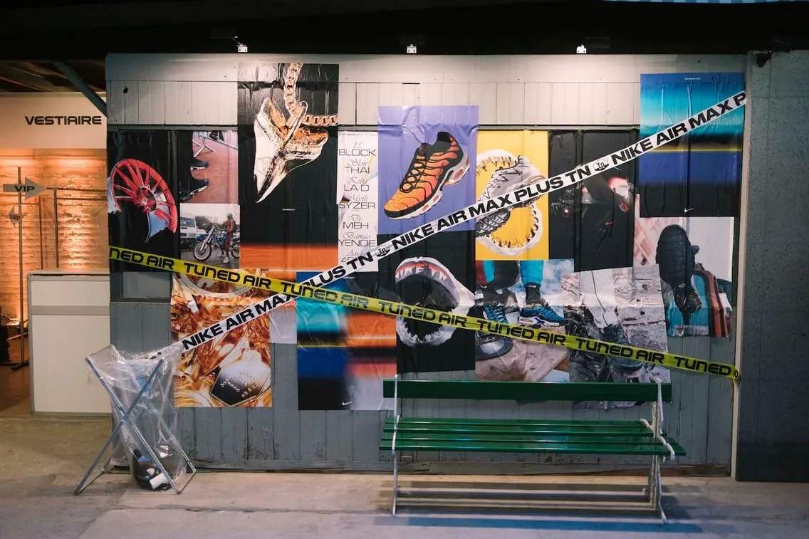 Mur d'affichage avec plusieurs affiches de chaussures Nike Air Max et Nike Air Max Plus TN, avec une bande jaune et blanche marqué Tuned Air et Nike Air Max Plus TN, devant un banc vert dans un espace intérieur industriel.