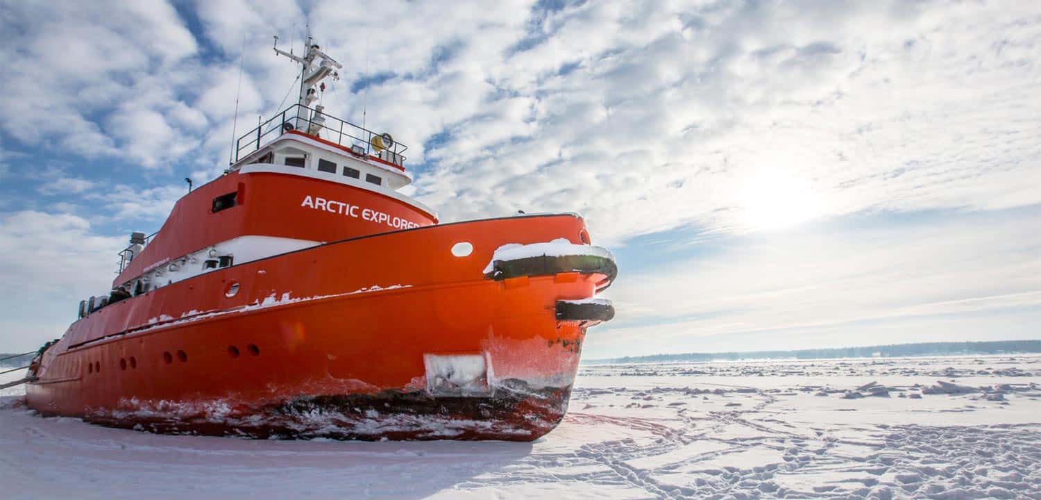 Orange icebreaker ship named Arctic Explorer navigating through icy, snow-covered waters under a partly cloudy sky.