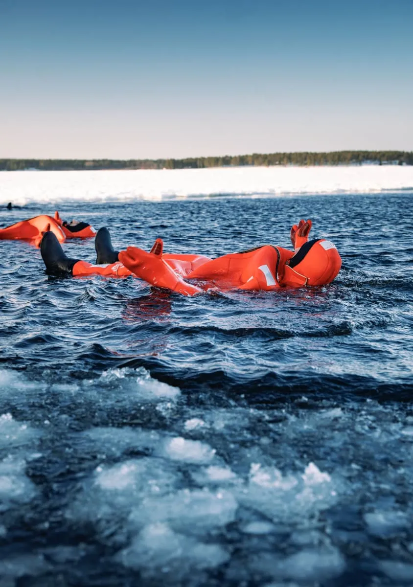 Two people in bright orange survival suits floating in icy water near a snowy shore under a clear blue sky.